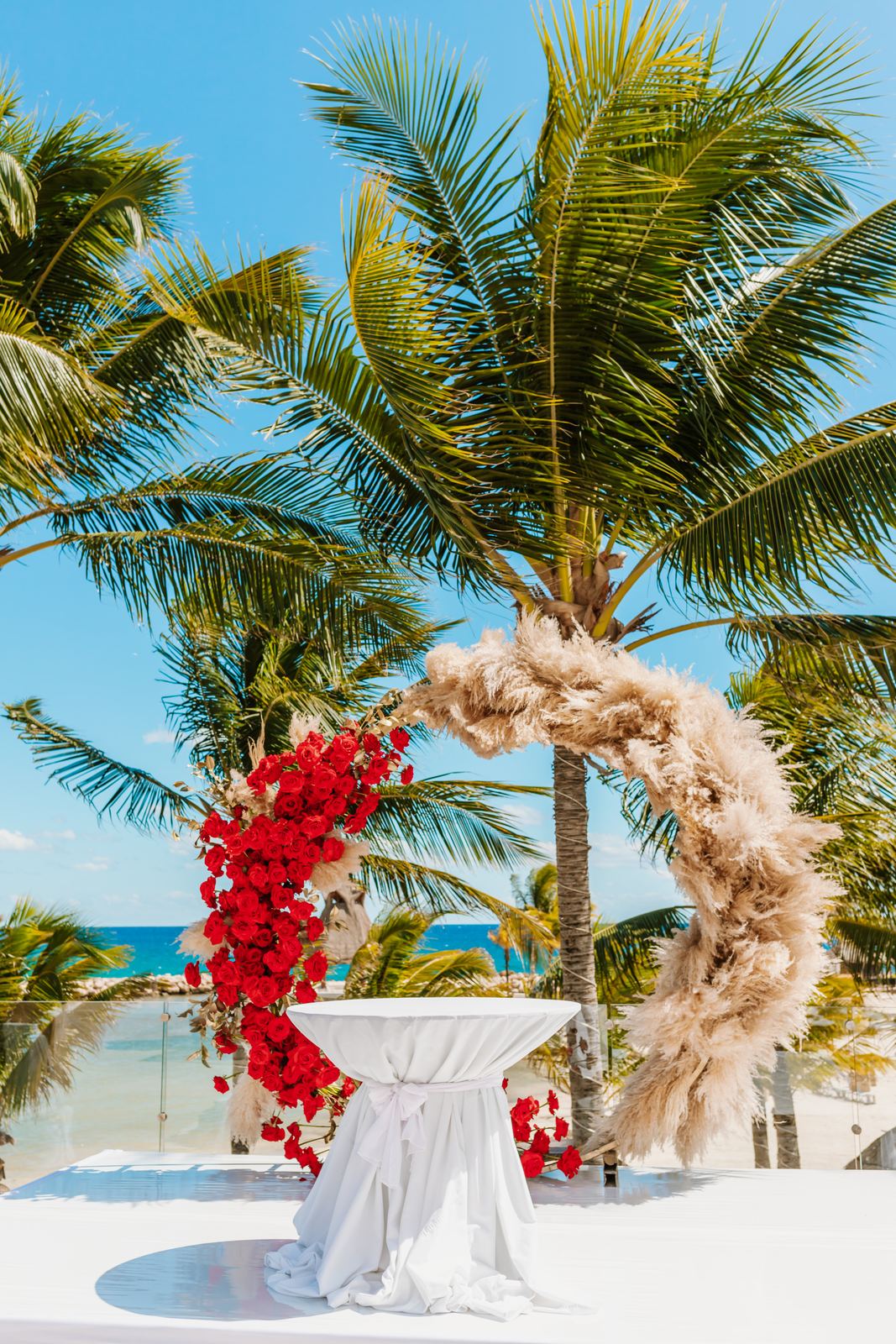 Red rose and pampas-grass ceremony arch under palms by the sea, set for the Catholic ceremony of a Hindu and Catholic wedding