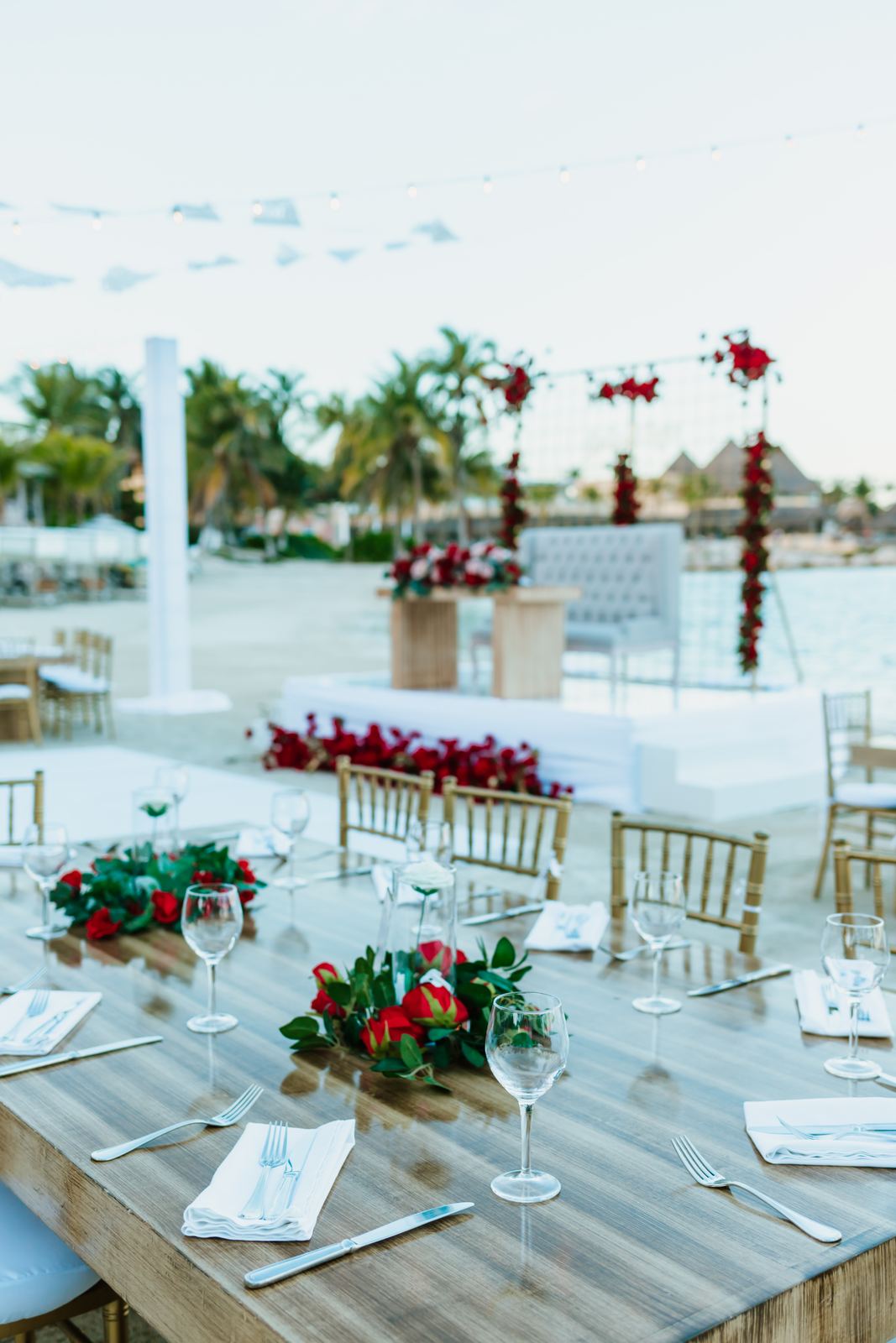 Reception table set with red roses by the sea, string lights over the sweetheart stage behind