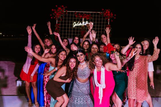 The bride and a crowd of guests, arms up, under the neon sign