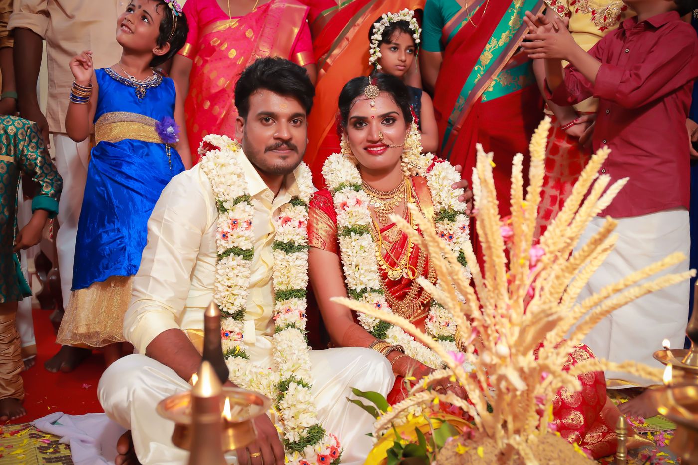 A Kerala couple seated behind brass nilavilakku lamps, garlanded in jasmine
