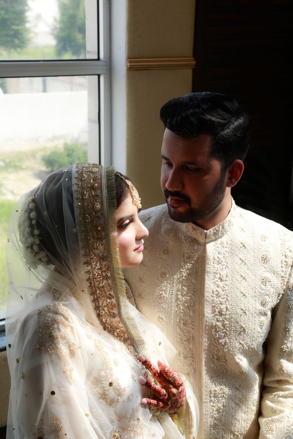 A bride in an ivory dupatta with mehndi beside her groom in a cream sherwani